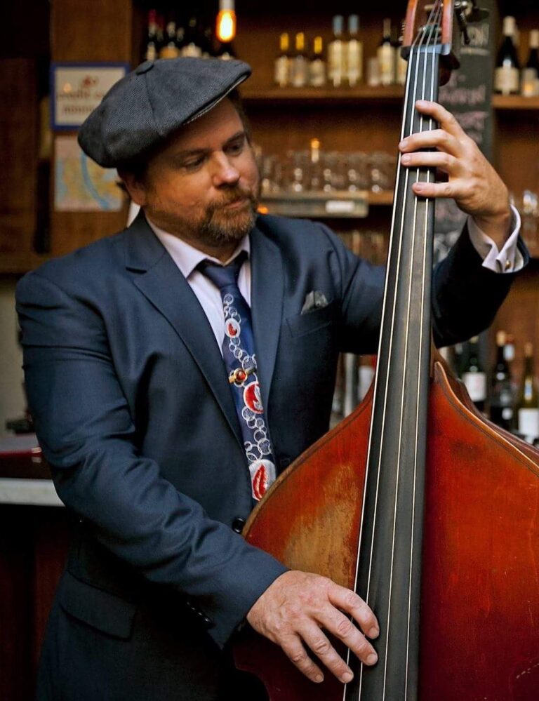 Joe Kyle Jr., a man with a cropped beard and mustache wearing a suit and hat, plays an upright bass.