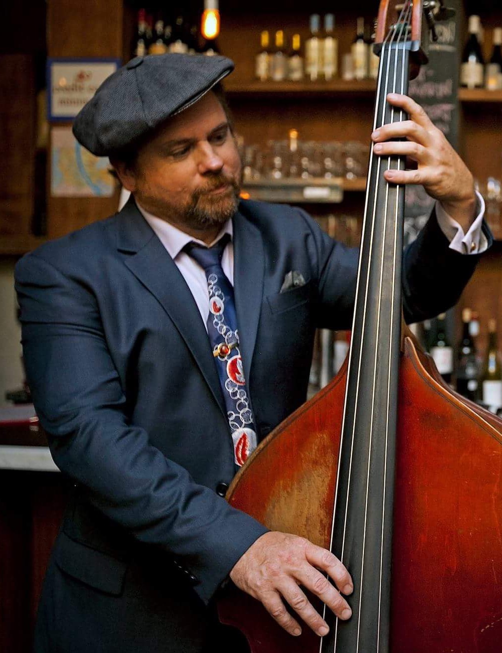 Joe Kyle Jr., a man with a cropped beard and mustache wearing a suit and hat, plays an upright bass.