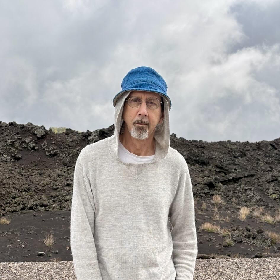 jd hanes, a man with a short mustache and beard wearing glasses, a blue hat, and light grey hoody, stands in front of a rocky hill.