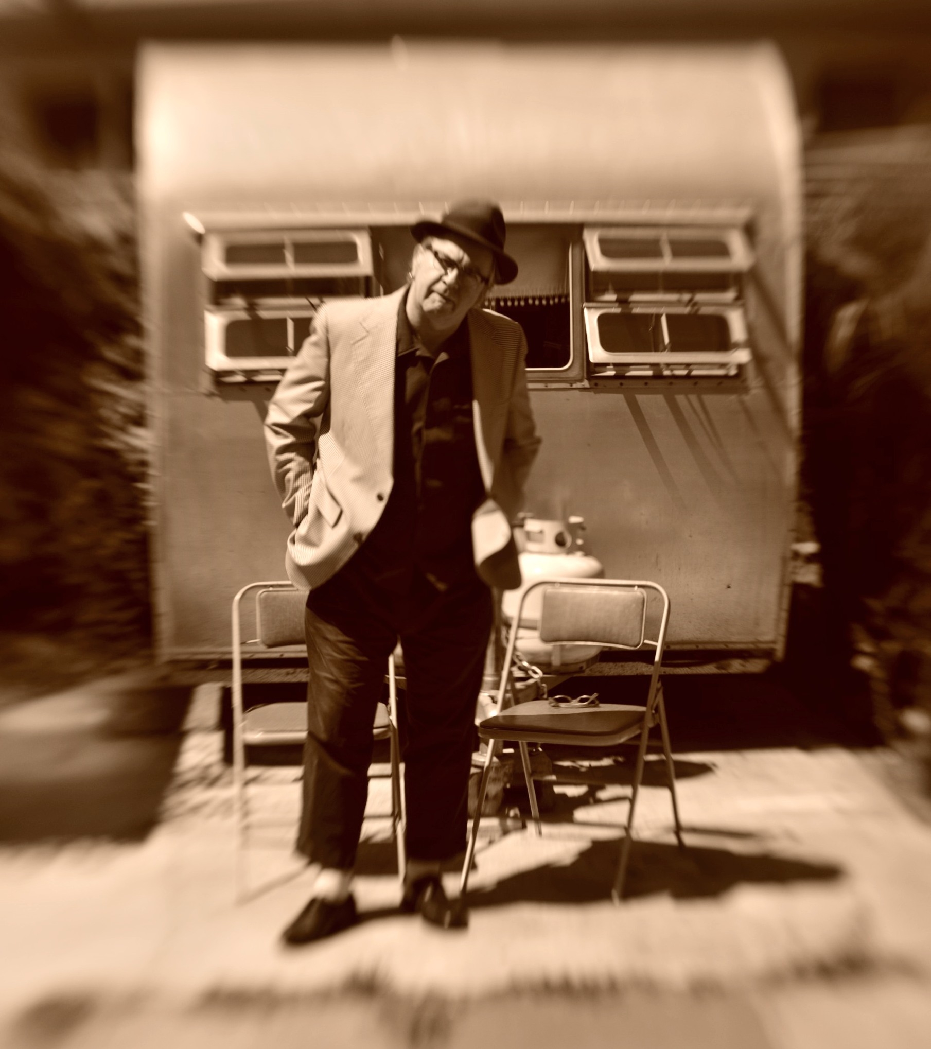 Myles Boisen, wearing a suit and hat, stands in front of a trailer. His hands are tucked into his jacket pockets. The edges of the photo are blurred.