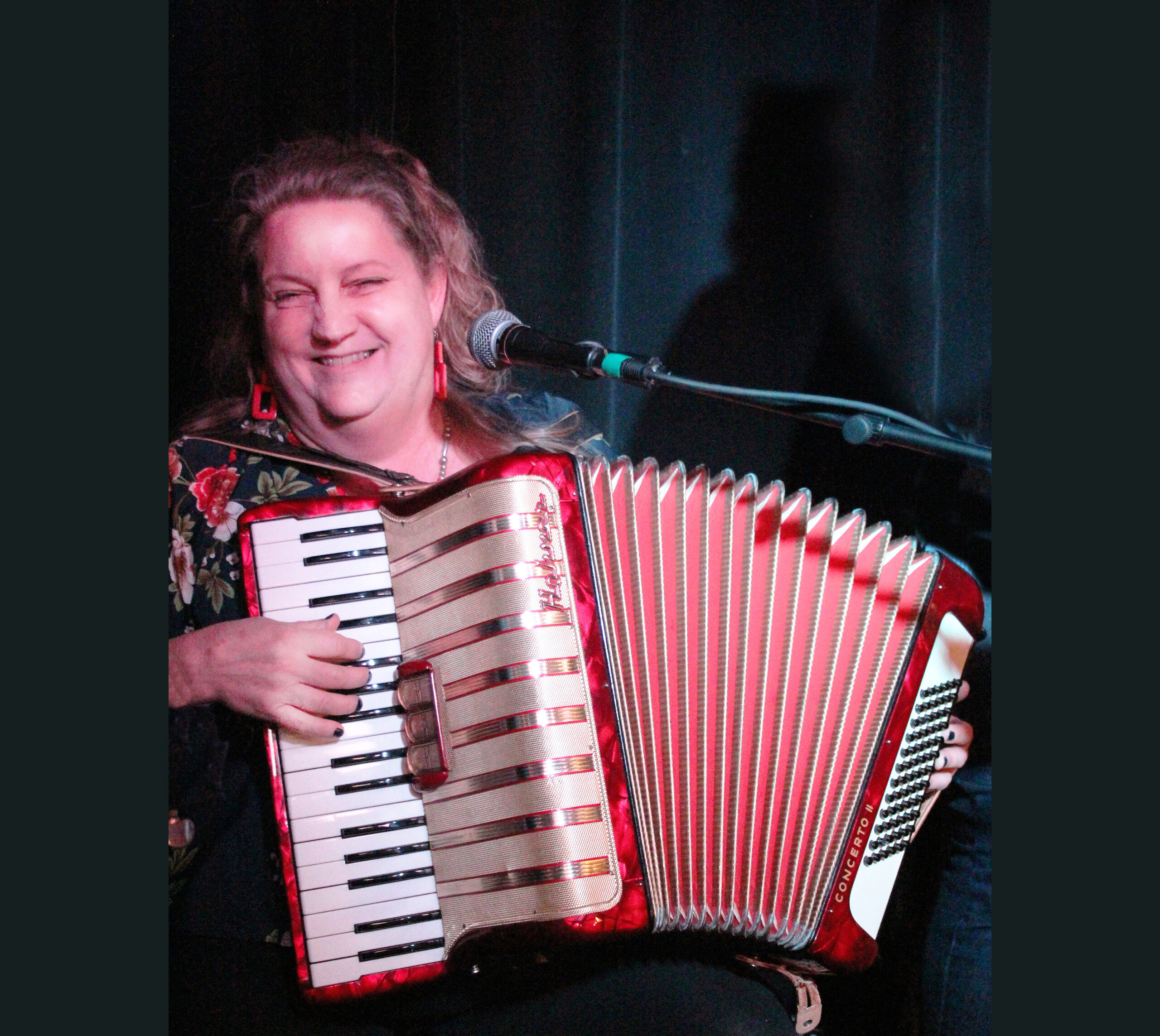 Sarah Gronquist, a woman wearing red earrings and a flower-print dress, plays accordion while smiling.