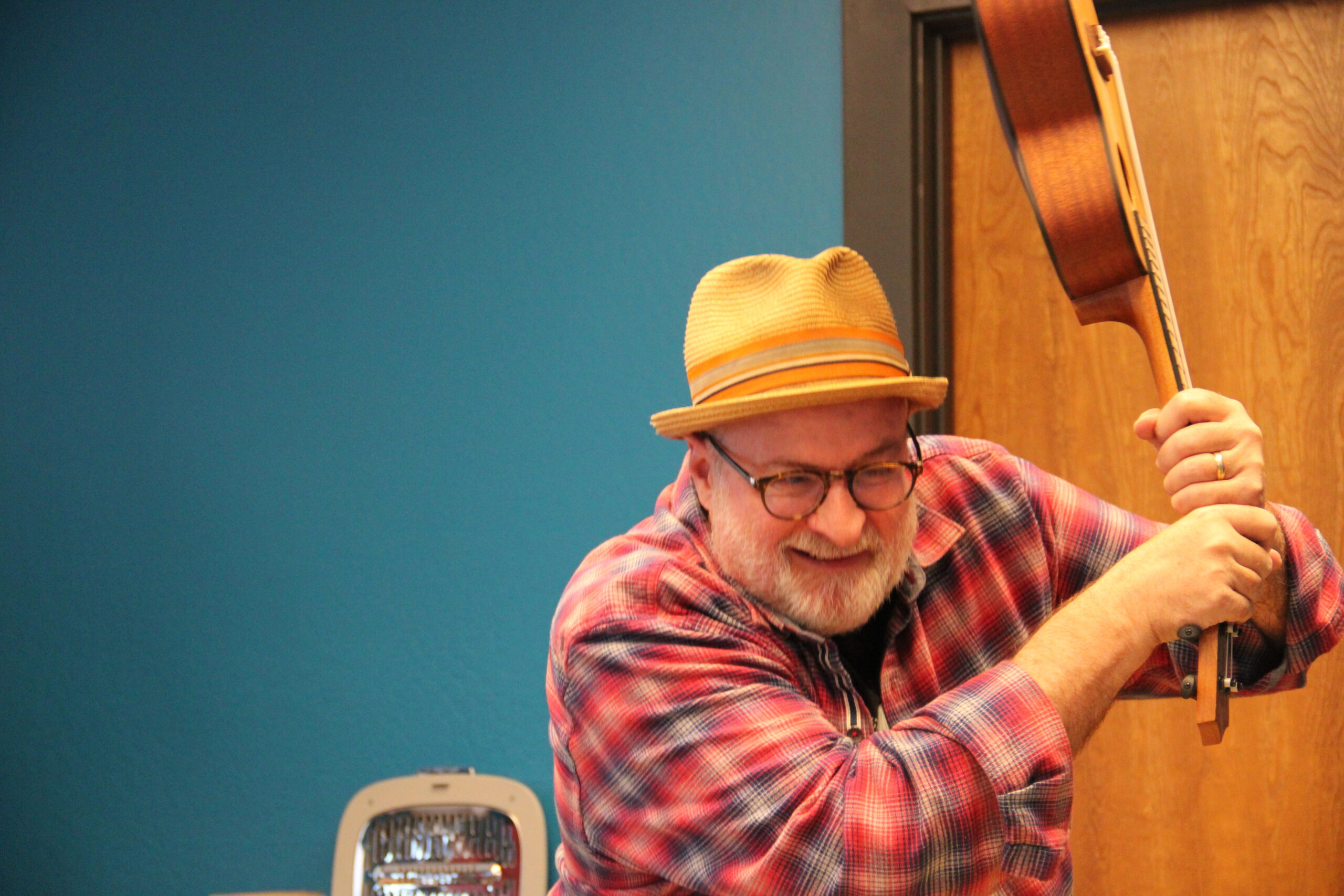 Christian Crumlish, a person with a beard wearing a red plaid shirt and yellow hat, raises a ukelele as if to smash it.
